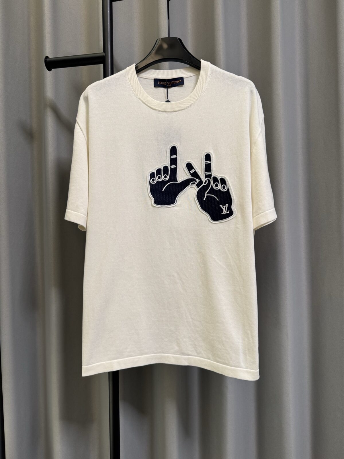 P155 【高端复刻】 𝐋𝐨𝐮𝐢𝐬 𝐕𝐮𝐢𝐭𝐭𝐨𝐧/路易 26SS lv手势刺绣针织短袖T恤（米白） LV 以趣味手势元素打造的创意系列短袖，用针织面料与立体刺绣工艺，打造兼具质感与个性的小众款。 🔹 面料与质感 核心面料：甄选LV 定制针织面料，布面细腻软糯，自带柔和哑光质感，上身轻薄透气，垂坠感优异，久穿不易起球变形。 穿着体验：面料亲肤透气，吸湿排汗性优异，夏季穿着清爽不闷汗；触感顺滑亲肤，不扎肤、不闷热，日常穿着舒适自在。 版型表现：合身直筒版型，修饰肩线与身形比例；圆领设计利落干练，自带简约高级氛围，适配多种穿搭场景。 🔹 工艺与细节 核心手势刺绣工艺： 胸口位置采用趣味手势刺绣，以藏蓝色为主体，搭配白色轮廓线，其中一只手上带有 LV 字母标识，刺绣针脚细密饱满，边缘无毛边，质感立体精致。在极简的米白色针织衣身上，这个创意手势刺绣是整件 T 恤的灵魂亮点。 细节做工： 领口采用罗纹收边，走线工整细密，反复穿着不易松垮变形； 袖口、下摆采用罗纹锁边，做工精细，不易脱线； 领标、洗水标等辅料齐全，完整还原正品配置，细节拉满。 Size：s/m/l/xl/
