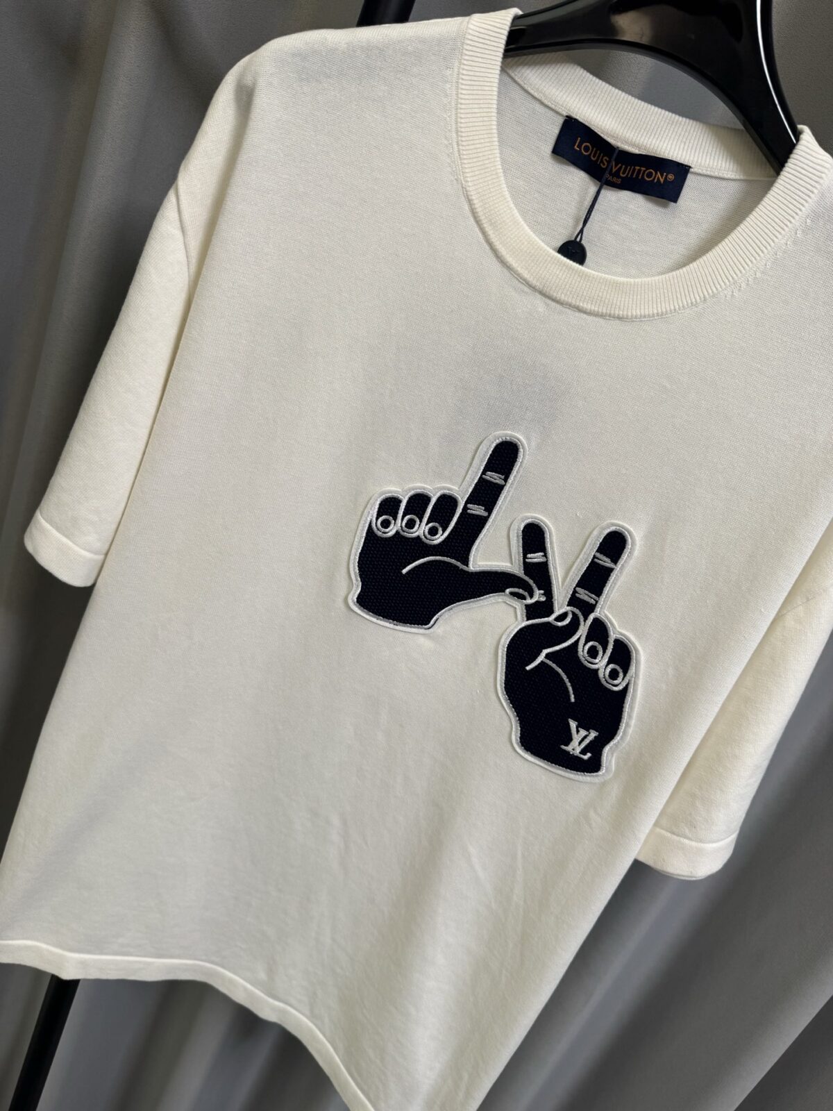 P155 【高端复刻】 𝐋𝐨𝐮𝐢𝐬 𝐕𝐮𝐢𝐭𝐭𝐨𝐧/路易 26SS lv手势刺绣针织短袖T恤（米白） LV 以趣味手势元素打造的创意系列短袖，用针织面料与立体刺绣工艺，打造兼具质感与个性的小众款。 🔹 面料与质感 核心面料：甄选LV 定制针织面料，布面细腻软糯，自带柔和哑光质感，上身轻薄透气，垂坠感优异，久穿不易起球变形。 穿着体验：面料亲肤透气，吸湿排汗性优异，夏季穿着清爽不闷汗；触感顺滑亲肤，不扎肤、不闷热，日常穿着舒适自在。 版型表现：合身直筒版型，修饰肩线与身形比例；圆领设计利落干练，自带简约高级氛围，适配多种穿搭场景。 🔹 工艺与细节 核心手势刺绣工艺： 胸口位置采用趣味手势刺绣，以藏蓝色为主体，搭配白色轮廓线，其中一只手上带有 LV 字母标识，刺绣针脚细密饱满，边缘无毛边，质感立体精致。在极简的米白色针织衣身上，这个创意手势刺绣是整件 T 恤的灵魂亮点。 细节做工： 领口采用罗纹收边，走线工整细密，反复穿着不易松垮变形； 袖口、下摆采用罗纹锁边，做工精细，不易脱线； 领标、洗水标等辅料齐全，完整还原正品配置，细节拉满。 Size：s/m/l/xl/