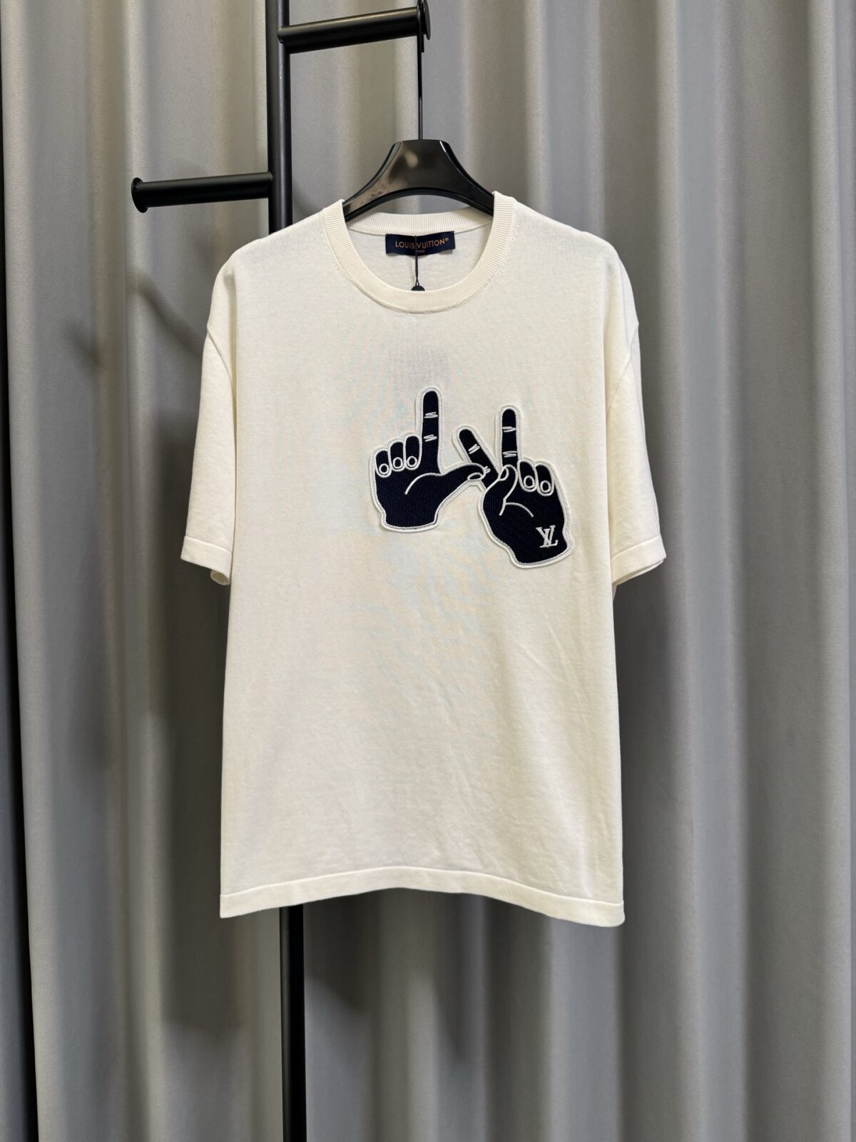 P155 【高端复刻】 𝐋𝐨𝐮𝐢𝐬 𝐕𝐮𝐢𝐭𝐭𝐨𝐧/路易 26SS lv手势刺绣针织短袖T恤（米白） LV 以趣味手势元素打造的创意系列短袖，用针织面料与立体刺绣工艺，打造兼具质感与个性的小众款。 🔹 面料与质感 核心面料：甄选LV 定制针织面料，布面细腻软糯，自带柔和哑光质感，上身轻薄透气，垂坠感优异，久穿不易起球变形。 穿着体验：面料亲肤透气，吸湿排汗性优异，夏季穿着清爽不闷汗；触感顺滑亲肤，不扎肤、不闷热，日常穿着舒适自在。 版型表现：合身直筒版型，修饰肩线与身形比例；圆领设计利落干练，自带简约高级氛围，适配多种穿搭场景。 🔹 工艺与细节 核心手势刺绣工艺： 胸口位置采用趣味手势刺绣，以藏蓝色为主体，搭配白色轮廓线，其中一只手上带有 LV 字母标识，刺绣针脚细密饱满，边缘无毛边，质感立体精致。在极简的米白色针织衣身上，这个创意手势刺绣是整件 T 恤的灵魂亮点。 细节做工： 领口采用罗纹收边，走线工整细密，反复穿着不易松垮变形； 袖口、下摆采用罗纹锁边，做工精细，不易脱线； 领标、洗水标等辅料齐全，完整还原正品配置，细节拉满。 Size：s/m/l/xl/