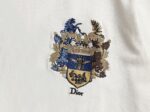 P80 迪奥 Dior密蜂皇冠徽章刺绣短袖T恤 采用100%精梳双股棉平纹针织面料织造，经预缩水洗工艺处理，布面平整光洁且抗缩率稳定，垂坠挺括兼具亲肤透气属性 廓形为Oversize落肩版型，圆领采用双针包边工艺，领口内置弹力罗纹，久洗不易变形。前中核心徽章图案采用多色立体盘绣工艺，以蓝、金、棕三色分层刺绣，通过长短针、打籽绣等针法塑造浮雕质感，将皇家纹章、骏马、鹰羽与品牌标志性蜜蜂元素精准呈现，绣线光泽度饱满且线迹密度均匀，边缘无跳线毛边 2026ss官方同步 高版本 高品质 时尚个性 百搭潮流 纯原品质 原版面料 吊牌 领标 水洗麦 三标齐全 颜色：白色 码数：S M L XL