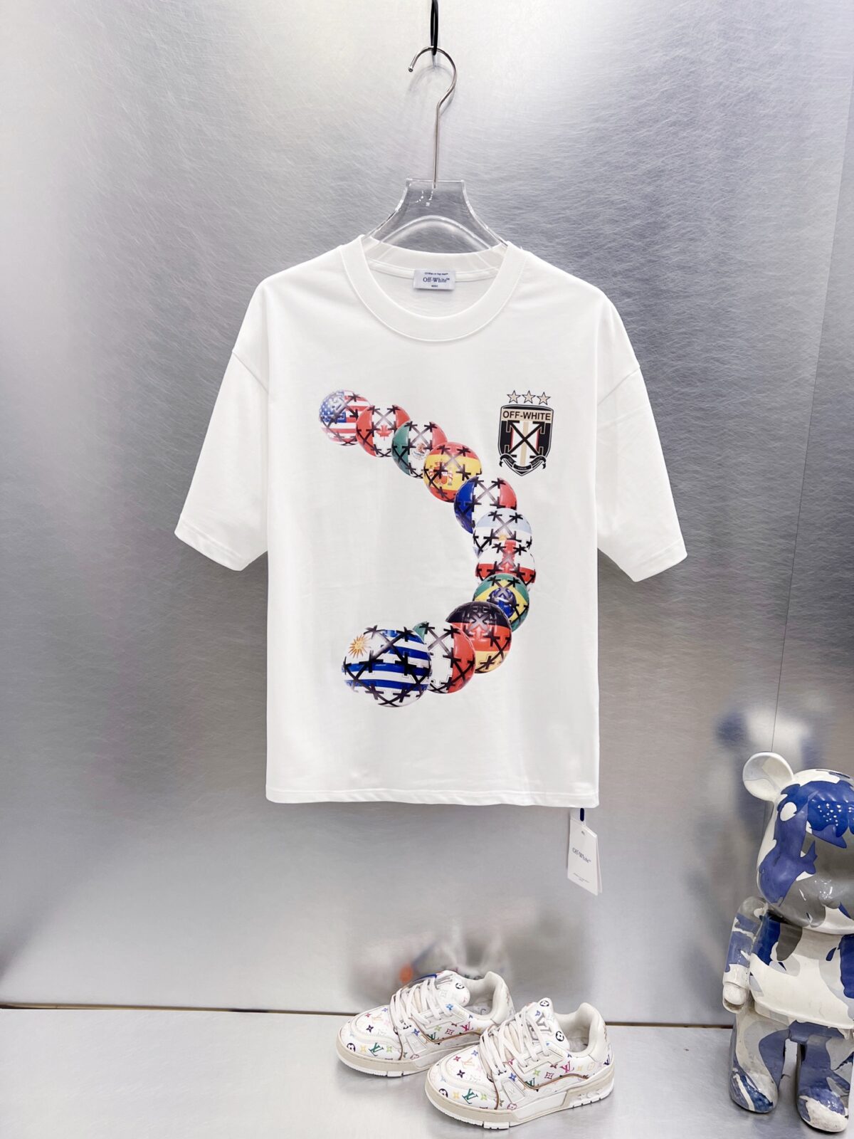 P75 OFF-WHITE 多国国旗足球印花短袖 世界杯徽章箭头重磅棉潮牌 T 恤，球迷潮人本命 OW 短袖来了！ 颜色：白色 尺码：S M L XL 整件衣服布满印满各国国旗的足球，每颗球都带有品牌标志性交叉箭头，侧边四星盾形徽章细节拉满，把世界杯环球氛围感直接穿身上。 重磅纯棉料子不透肉，宽松版型藏肉显瘦，看球约会日常穿都合适，喜欢足球和街头风的男生女生直接冲，情侣搭一套超吸睛！