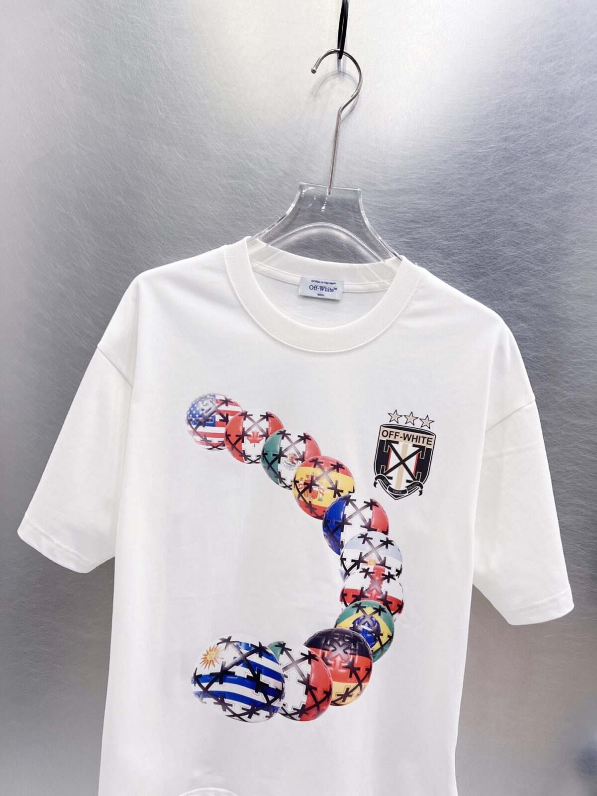 P75 OFF-WHITE 多国国旗足球印花短袖 世界杯徽章箭头重磅棉潮牌 T 恤，球迷潮人本命 OW 短袖来了！ 颜色：白色 尺码：S M L XL 整件衣服布满印满各国国旗的足球，每颗球都带有品牌标志性交叉箭头，侧边四星盾形徽章细节拉满，把世界杯环球氛围感直接穿身上。 重磅纯棉料子不透肉，宽松版型藏肉显瘦，看球约会日常穿都合适，喜欢足球和街头风的男生女生直接冲，情侣搭一套超吸睛！