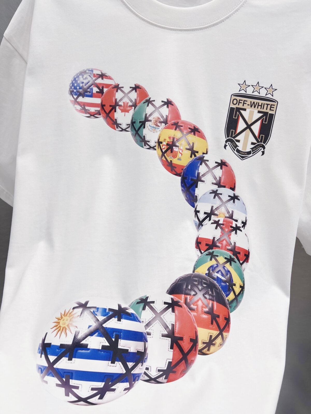 P75 OFF-WHITE 多国国旗足球印花短袖 世界杯徽章箭头重磅棉潮牌 T 恤，球迷潮人本命 OW 短袖来了！ 颜色：白色 尺码：S M L XL 整件衣服布满印满各国国旗的足球，每颗球都带有品牌标志性交叉箭头，侧边四星盾形徽章细节拉满，把世界杯环球氛围感直接穿身上。 重磅纯棉料子不透肉，宽松版型藏肉显瘦，看球约会日常穿都合适，喜欢足球和街头风的男生女生直接冲，情侣搭一套超吸睛！